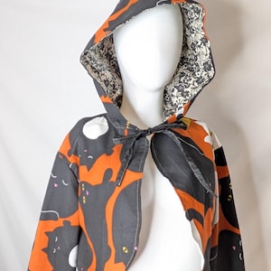 May include: A cropped hooded cape featuring a black and orange cat pattern on the exterior and a black and white floral print lining. The garment includes a pointed hood and a fabric tie closure at the neck, worn over a matching elastic waist skirt.