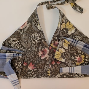 Puede incluir: Top halter con estampado floral en tonos marrón, rosa y amarillo. El top tiene lazos y bordes a rayas azules y blancas. La tela presenta un diseño botánico con hojas y flores.