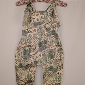 May include: A baby jumpsuit with a patchwork design in shades of green, beige, and gray, hanging on a floral-patterned hanger. The jumpsuit features a variety of geometric and floral patterns. The straps are tied at the shoulders.