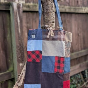 May include: A patchwork tote bag made from blue denim, red and black plaid, and brown fabric. The bag has a blue denim strap and a small brown leather tag with the word "BVE" on it.