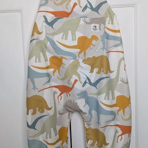 Peut inclure: Salopette pour bébé à motif de dinosaures aux couleurs douces. Le vêtement présente un motif répété de dinosaures orange, jaune, bleu et vert. La salopette est suspendue à un cintre bleu et en bois.