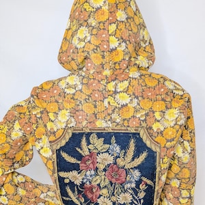 May include: A hooded jacket featuring an all-over floral pattern in shades of yellow, orange, and brown. The back displays a large, rectangular dark blue patch with a detailed embroidered bouquet of red poppies, wheat, and white flowers.