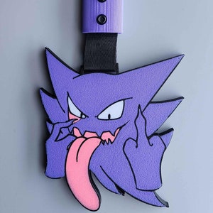May include: A purple cartoon character with a pink tongue and a black strap. The character is giving the middle finger. The strap is black and attached to a purple plastic piece with two black rivets.