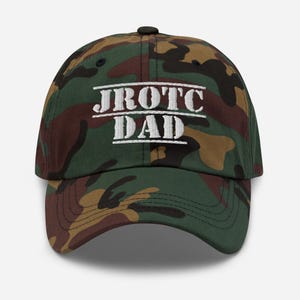 May include: A camouflage baseball cap with the text "JROTC DAD" in white block letters. The cap features a curved brim and a black button on top. The camouflage pattern includes shades of green, brown, and tan.