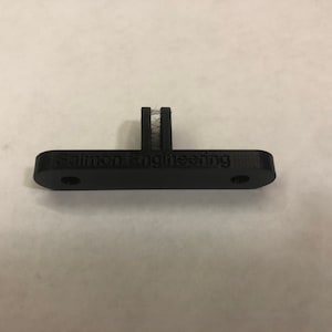 Peut inclure: Une pièce en plastique noire imprimée en 3D avec le texte "Salmon Engineering" imprimé dessus. La pièce a une petite pièce en plastique blanche fixée sur le dessus.