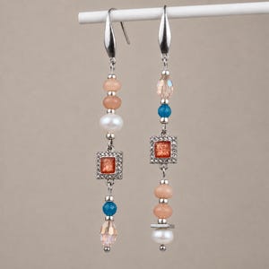 Könnte beinhalten: Ein Paar lange Ohrhänger mit silberfarbenen Haken, pfirsichfarbenen runden Perlen, weißen Perlen, kleinen blauen Perlen und quadratischen Anhängern mit orangefarbenem Glitzerkern, die von winzigen klaren Kristallen umrahmt sind.