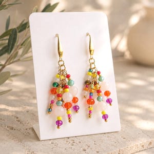 Könnte beinhalten: Ein Paar goldfarbene Ohrhänger mit einem mehrsträngigen Quasten-Design. Jeder Ohrring ist mit einer Auswahl an kleinen, bunten Glas- und Steinperlen in den Farben Pink, Gelb, Orange, Grün, Blau und Violett verziert, die an Goldringen hängen.