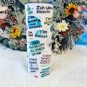 May include: White stainless steel tumbler with a lid, featuring handwritten affirmations and colorful abstract shapes. Text includes phrases like "I am powerful and confident" and "I am enough". A motivational and inspirational item.