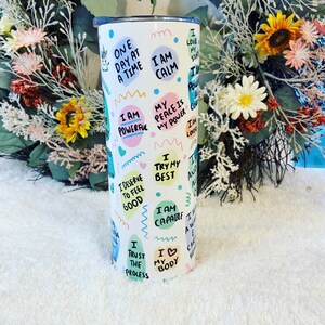 May include: White tumbler with a lid, covered in colorful text and illustrations. Phrases like "I am powerful," "I am calm," and "I love my story" are featured. A unicorn graphic is also present. The tumbler is cylindrical.