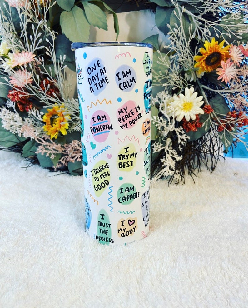 May include: White tumbler with a lid, covered in colorful text and illustrations. Phrases like "I am powerful," "I am calm," and "I love my story" are featured. A unicorn graphic is also present. The tumbler is cylindrical.