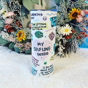 May include: A white tumbler with a blue lid, featuring colorful text and illustrations. The tumbler has phrases like "My Self Love Guide" and "I am the healer of my own life."