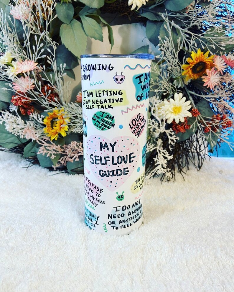May include: A white tumbler with a blue lid, featuring colorful text and illustrations. The tumbler has phrases like "My Self Love Guide" and "I am the healer of my own life."