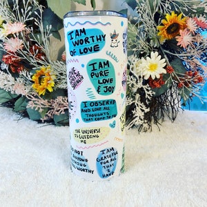 May include: A white stainless steel tumbler with a colorful design featuring affirmations like "I am worthy of love" and "I am pure love & joy." The tumbler has a unicorn graphic and various text bubbles with positive messages.