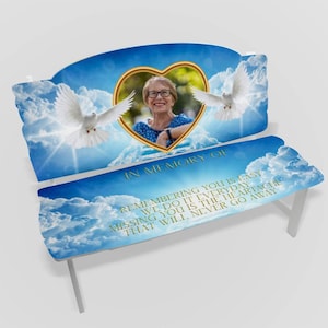 May include: White wooden bench with a blue sky and clouds design. A gold heart with a photo of a woman is in the centre of the bench. The text "In Memory Of" and a quote about remembering someone are printed on the bench.