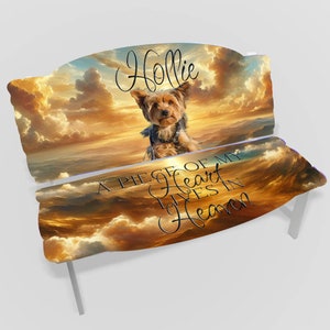 May include: A white wooden bench with a sunset sky and clouds design. The bench has a personalized inscription that reads "Hollie, A piece of my heart lives in Heaven". A small Yorkshire Terrier dog is sitting on the bench.