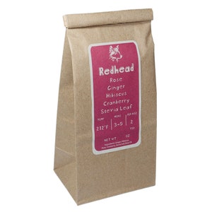 Puede incluir: Una bolsa de papel marrón con una etiqueta rosa que dice "Redhead" y enumera los ingredientes: Rosa, Jengibre, Hibisco, Arándano, Stevia Leaf. La etiqueta también incluye instrucciones de preparación: 212°F, 3-5 minutos, 2 cucharaditas por 2 oz.