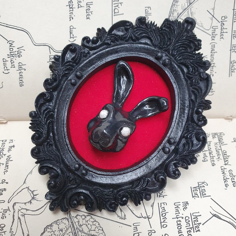 May include: A black rabbit figurine with white eyes is displayed in a black ornate frame with a red velvet background.