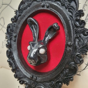 May include: A black rabbit sculpture with white eyes is displayed in a black ornate frame with a red velvet background.