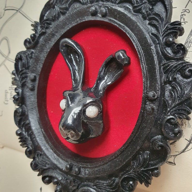 May include: A black rabbit sculpture with white eyes is displayed in a black ornate frame with a red velvet background.