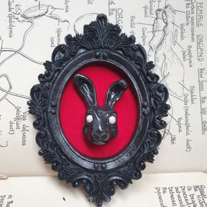 May include: A black rabbit sculpture with white eyes, framed in a black ornate frame with a red velvet background.