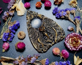 May include: A gold-colored, ornate, triangular-shaped planchette with a circular window in the center. The planchette is surrounded by dried flowers and other natural elements.