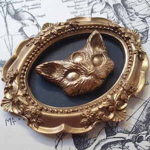 May include: A gold-colored cat head sculpture with three eyes is displayed in a gold ornate frame with a dark blue background.