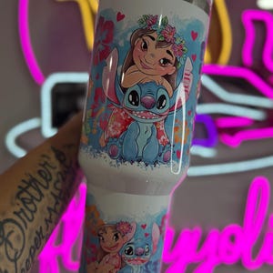 Puede incluir: Un vaso de acero inoxidable blanco con un dise&ntilde;o de dibujos animados colorido de Lilo y Stitch. El dise&ntilde;o incluye detalles florales y corazones. El vaso tiene una tapa plateada y mide aproximadamente 887 ml.