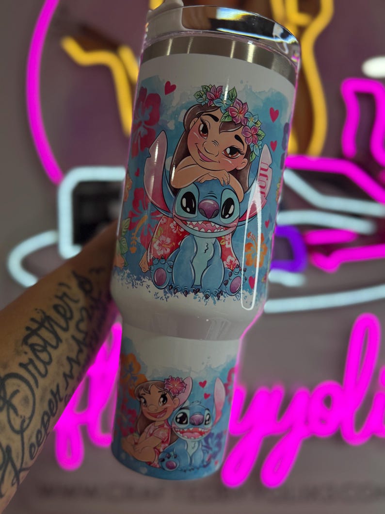 Puede incluir: Un vaso de acero inoxidable blanco con un dise&ntilde;o de dibujos animados colorido de Lilo y Stitch. El dise&ntilde;o incluye detalles florales y corazones. El vaso tiene una tapa plateada y mide aproximadamente 887 ml.