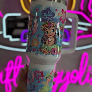 Puede incluir: Un vaso t&eacute;rmico blanco con asa, con un dise&ntilde;o colorido de una ni&ntilde;a y un extraterrestre azul con detalles florales. El dise&ntilde;o incluye corazones y flores. El vaso tiene una tapa blanca.