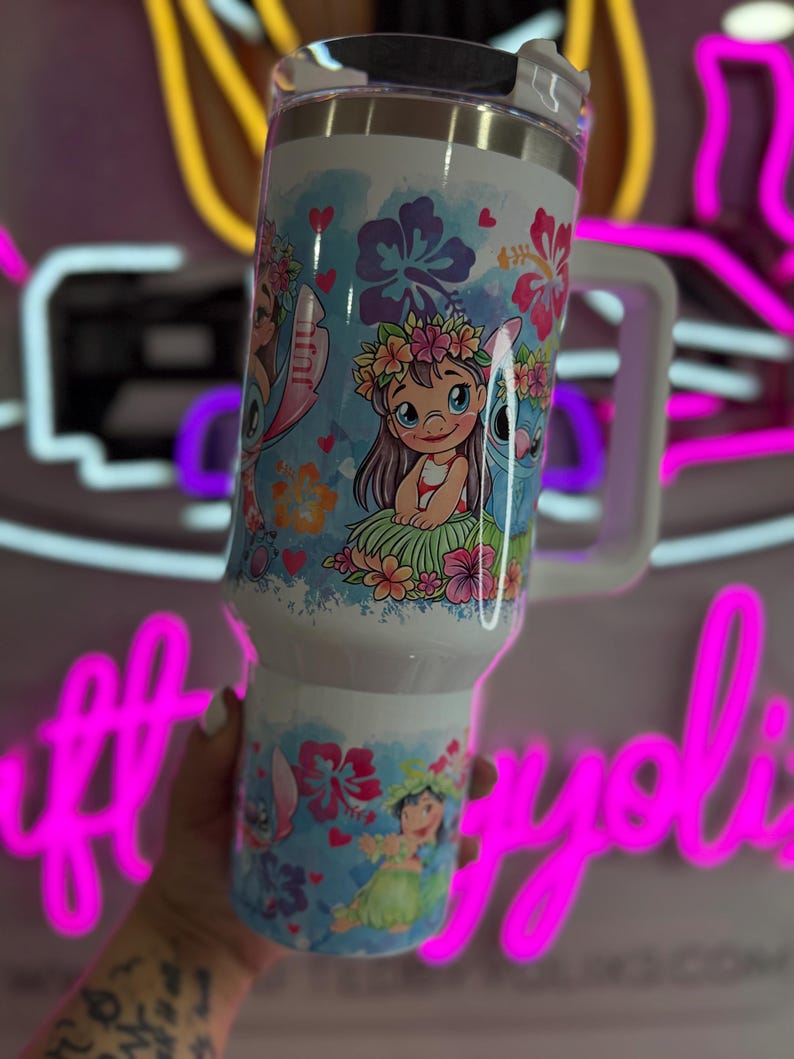 Puede incluir: Un vaso t&eacute;rmico blanco con asa, con un dise&ntilde;o colorido de una ni&ntilde;a y un extraterrestre azul con detalles florales. El dise&ntilde;o incluye corazones y flores. El vaso tiene una tapa blanca.