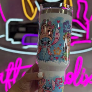 Puede incluir: Un vaso blanco con los personajes de dibujos animados Stitch y Moana, decorado con dise&ntilde;os florales y corazones. El vaso tiene una tapa transparente. El dise&ntilde;o est&aacute; sobre un fondo blanco.