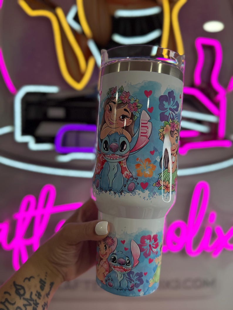 Puede incluir: Un vaso blanco con los personajes de dibujos animados Stitch y Moana, decorado con dise&ntilde;os florales y corazones. El vaso tiene una tapa transparente. El dise&ntilde;o est&aacute; sobre un fondo blanco.