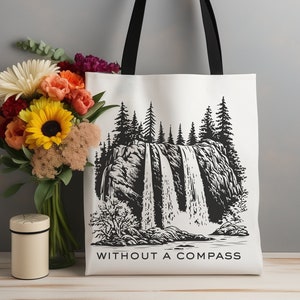 May include: A white tote bag with black handles and a black and white illustration of a waterfall in a forest. The text "WITHOUT A COMPASS" is printed on the bottom of the bag.