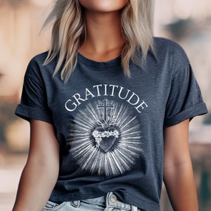Może przedstawiać: Ciemnoszary melanżowy t-shirt z kr&oacute;tkim rękawem z białym nadrukiem graficznym przedstawiającym Najświętsze Serce otoczone koroną cierniową, zwieńczone krzyżem i promieniami światła, z napisem GRATITUDE wygiętym w łuk nad wzorem.