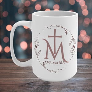 May include: White ceramic mug with a floral wreath design featuring a brown "Ave Maria" inscription and a stylised "M" with a cross.