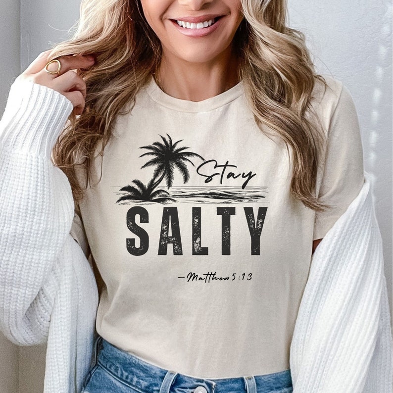 K&ouml;nnte beinhalten: Ein beiges T-Shirt mit einer schwarzen Grafik von Palmen und Wellen. Der Text "Stay Salty" ist in Schwarz mit einer verwitterten Schrift gedruckt. Der Text "Matthew 5:13" ist in einer kleineren Schrift unterhalb der Grafik gedruckt.