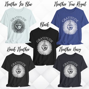Może przedstawiać: Pięć koszulek z kr&oacute;tkim rękawem w kolorach heather ice blue, heather true royal, czarnym, dark heather i heather navy. Każda koszulka posiada białą grafikę serca z krzyżem i promieniami oraz nadrukowane łukiem słowo GRATITUDE powyżej.