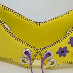 Puede incluir: Un accesorio de cuero amarillo brillante con un borde cosido y detalles morados. Cuenta con ojales plateados, cordones morados y beige, y adornos florales morados y amarillos. El diseño es simétrico y tiene una forma única.