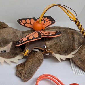 May include: A plush toy lizard with a butterfly design. The brown lizard has a cream-coloured belly and a butterfly with orange and black wings. It has a yellow and brown braided leash and orange cord.