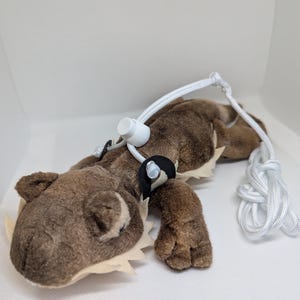 Op de afbeelding: Een bruin pluche dinosaurus speelgoed met een witte touwriem en een wit plastic stuk. De dinosaurus heeft een beige buik en een beige kraag.