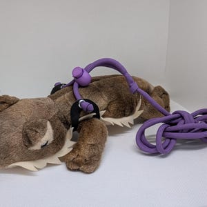 May include: A brown plush toy with a purple leash and harness. The toy has a light tan belly and a white, jagged trim. The purple leash is knotted and has a purple handle. The toy is lying on a white surface.