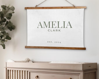 May include: A white canvas wall hanging with the text "Amelia Clark Est. 2024" in green lettering. The hanging is displayed above a light wood dresser with three drawers and a crib.