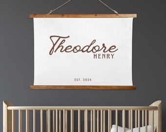 May include: A white canvas with a wooden frame, hanging on a gray wall. The canvas features the name "Theodore Henry" in brown cursive script, with the date "EST. 2024" below it. A crib with a white mattress and yellow pillow is in the foreground.