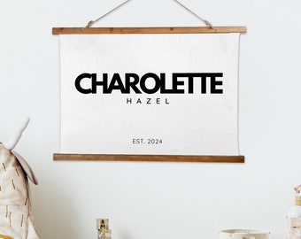 May include: A white canvas print with black text that reads "CHARLOTTE HAZEL EST. 2024". The print is hanging on a white wall in a nursery with a stuffed animal, wooden blocks, and a woven storage basket.