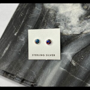 May include: A pair of sterling silver stud earrings with round, iridescent opal stones. The earrings are displayed on a white card with the text "STERLING SILVER".