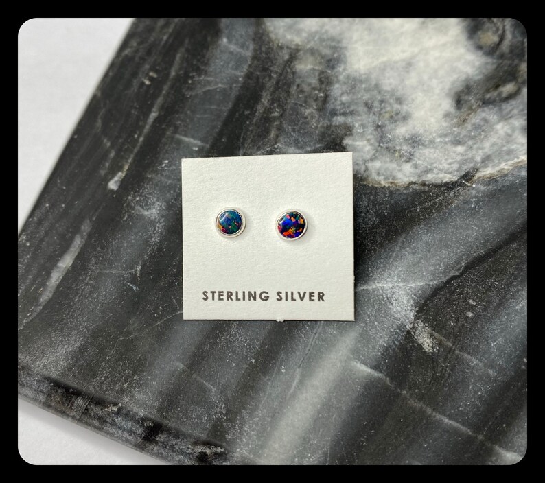May include: A pair of sterling silver stud earrings with round, iridescent opal stones. The earrings are displayed on a white card with the text "STERLING SILVER".