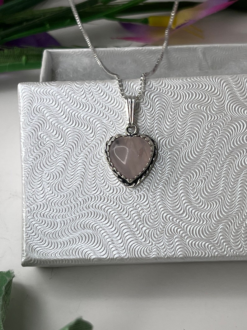 Puede incluir: Un collar de cadena de plata con un colgante en forma de coraz&oacute;n con una piedra de cuarzo rosa. El colgante tiene un delicado borde de plata.