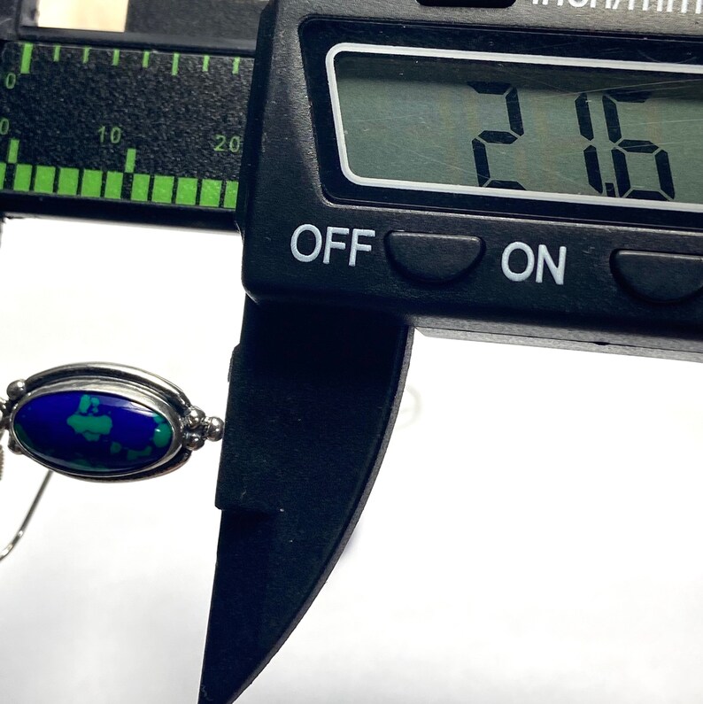 May include: Close-up of a blue and green gemstone pendant measured with a digital caliper reading 2.06. The pendant is oval shaped and set in silver.