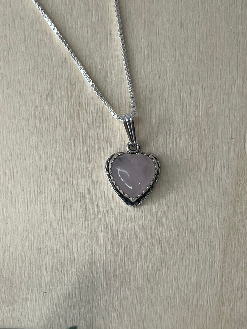 Puede incluir: Un collar de cadena de plata con un colgante en forma de coraz&oacute;n con una piedra preciosa rosa p&aacute;lida.