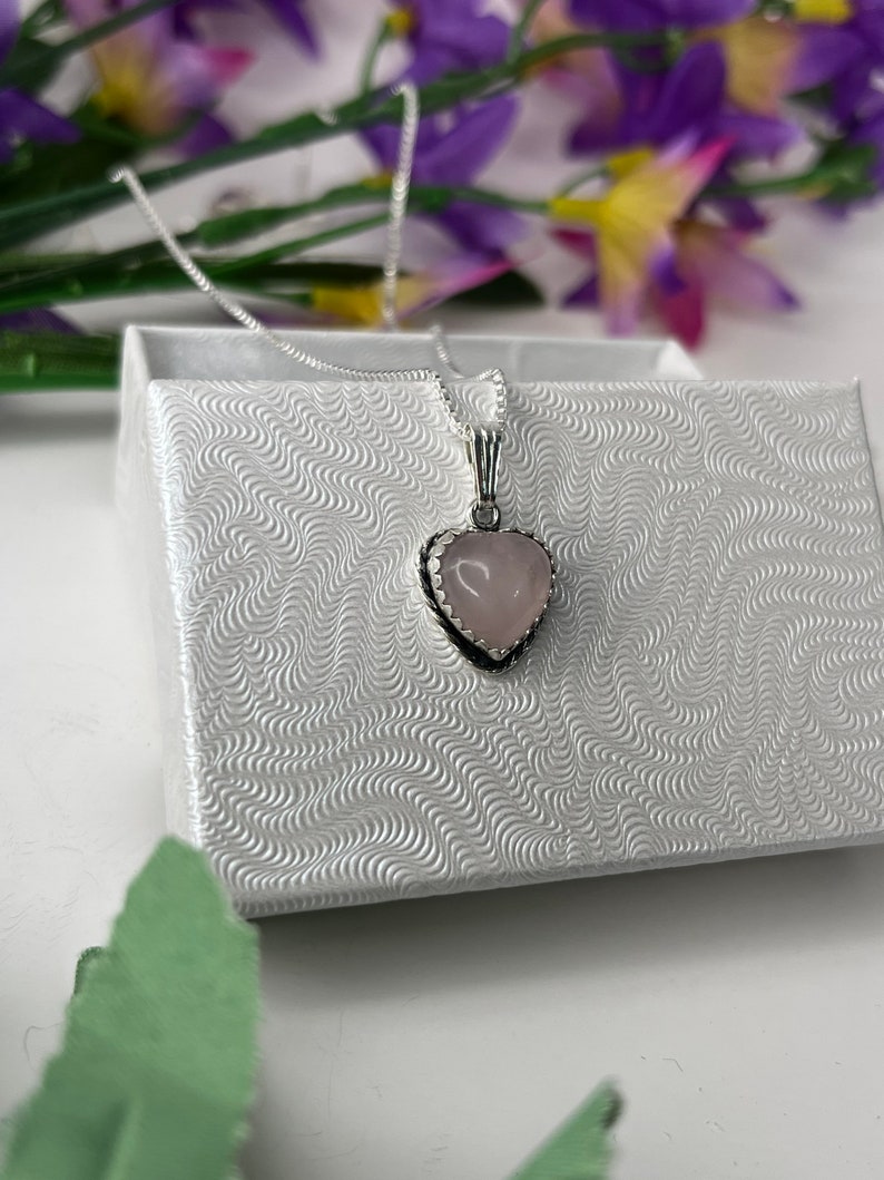 Puede incluir: Un collar de cadena de plata con un colgante de coraz&oacute;n rosa. El colgante tiene un borde plateado y un dise&ntilde;o delicado.
